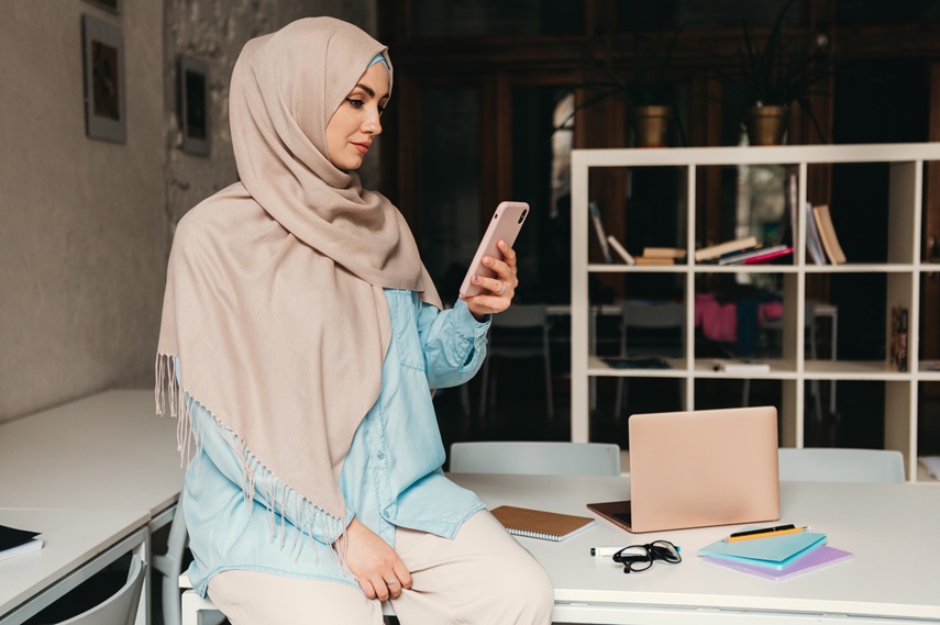 young pretty modern muslim woman in hijab working in office room, education online, remote work freelancer, using smartphone