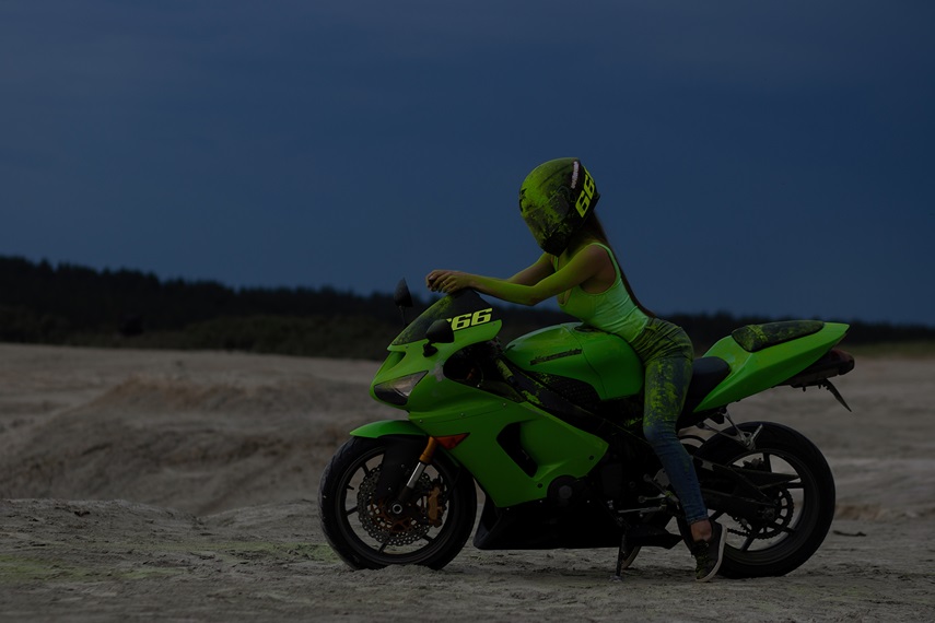 Anonymous cool female motorcyclist in helmet and green Holi powder sitting on contemporary motorbike on beach at sundown