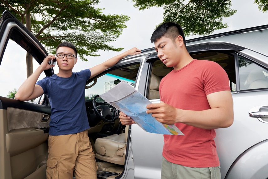 Taking rests after long road trip: young Asian man in eyeglasses using smartphone while his concentrated friend reading paper map